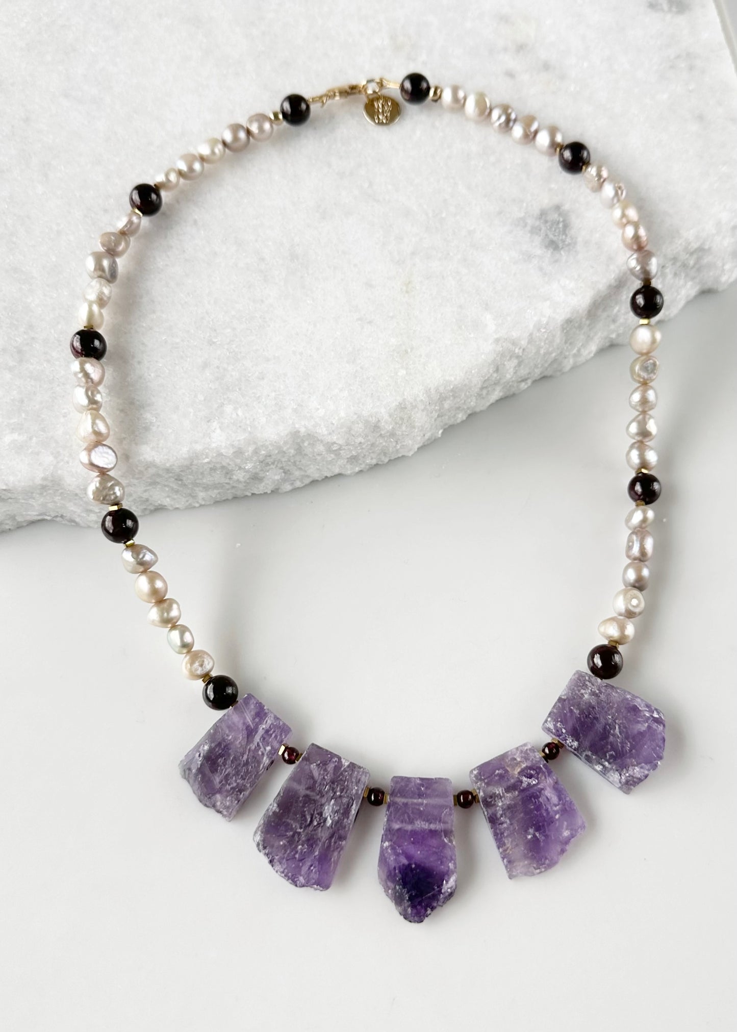 Designed and made in Canada, this Striking and Unique 20.5” Necklace showcases a set of 5 Natural Amethyst Pendants, with Baroque Champagne Pearls and pretty Round Garnets.  This piece is finished with a Gold-filled Lobster Claw Clasp.