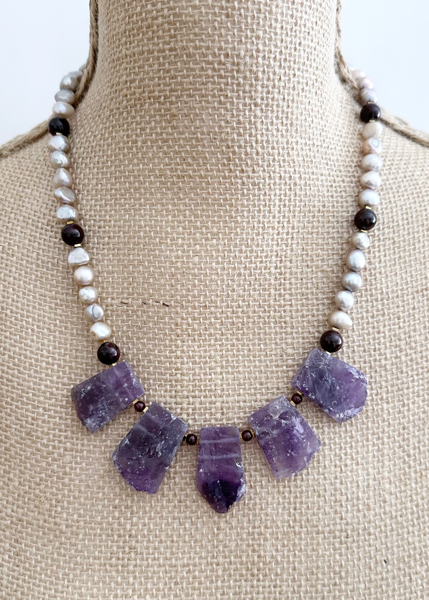Designed and made in Canada, this Striking and Unique 20.5” Necklace showcases a set of 5 Natural Amethyst Pendants, with Baroque Champagne Pearls and pretty Round Garnets.  This piece is finished with a Gold-filled Lobster Claw Clasp.