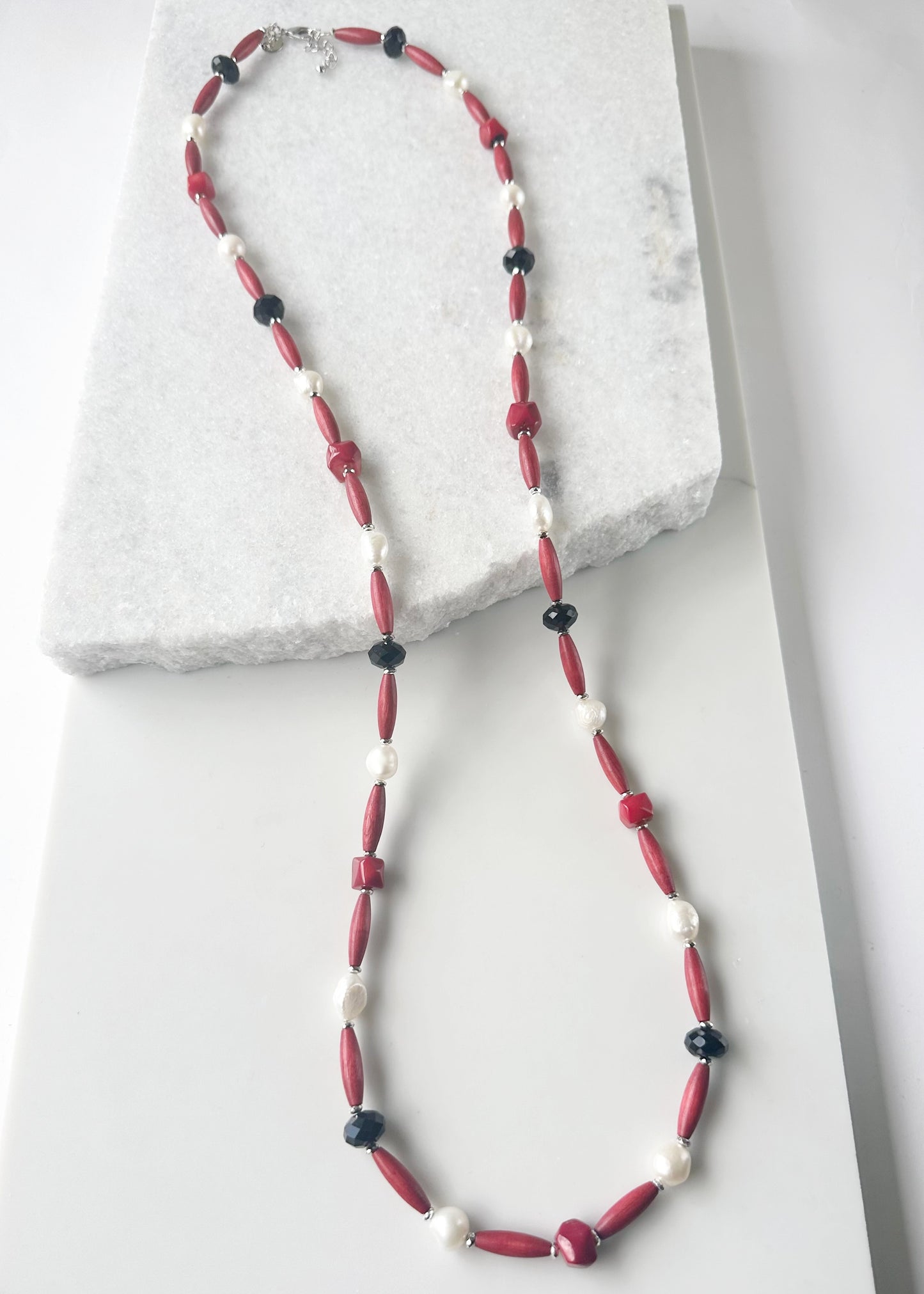 Designed and Made in Canada, this One-Of-A-Kind 40” Necklace Combines Red Bamboo Coral Nuggets, Faceted Black Crystals, Warm Red Wood, and Lustrous White Freshwater Pearls. The piece is Accented with Silver Hematite for depth and Finished with a Lobster Claw Clasp and 1.5