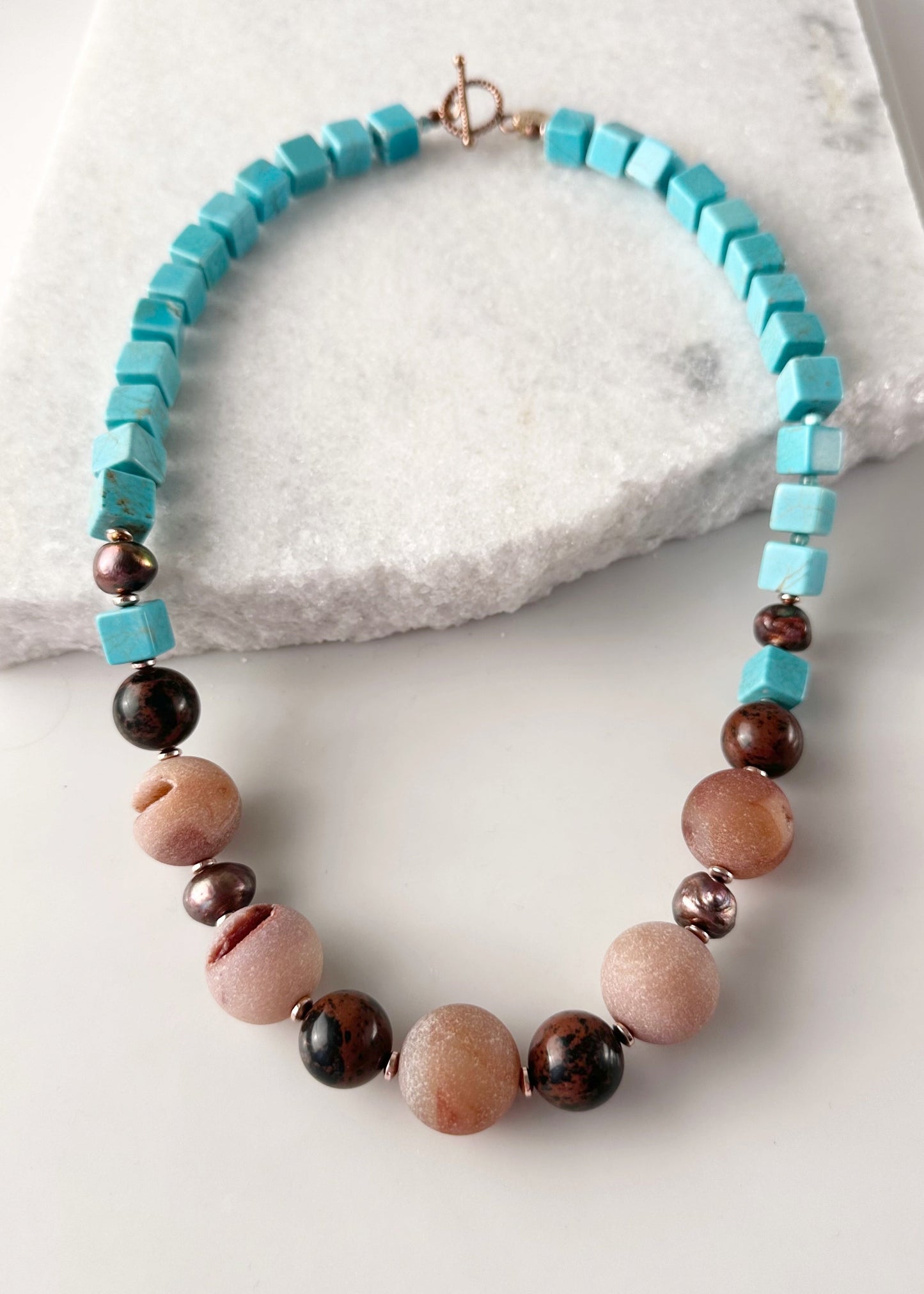 Designed and made in Canada, this One-Of-A-Kind 24” Necklace features Large Round Druzy Agate, Mahogany Obsidian, Turquoise Magnesite and Beautiful Brown Pearls. This piece is completed with Glass Seed Beads, Rose Gold-Plated Hematite and a Copper Toggle Clasp.