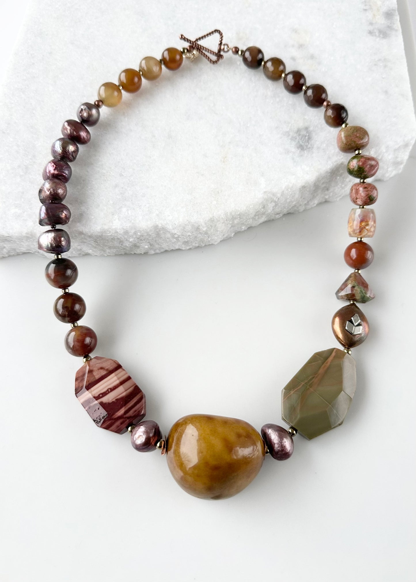 Designed and made in Canada, this Stunning One-Of-A-Kind 22.5" Necklace Showcases an Exquisite Blend of Imperial Jasper, Tagua Seed, Petrified Wood, Opalite, Unakite, and Agate, Accented with Lustrous Brown Pearls and Warm Copper and Gold details. Each element is thoughtfully combined to create a truly distinctive accessory that celebrates nature's palette and timeless elegance.