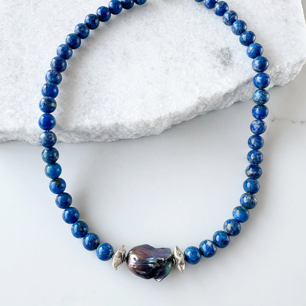
                      
                        A Large Peacock Baroque Freshwater Pearl sits Center Stage amidst Silver Accents and Beautiful Blue Lapis Lazuli Beads on this One-Of-A-Kind 20” Necklace.  It is finished with a Sterling Silver Lobster Claw Clasp.
                      
                    