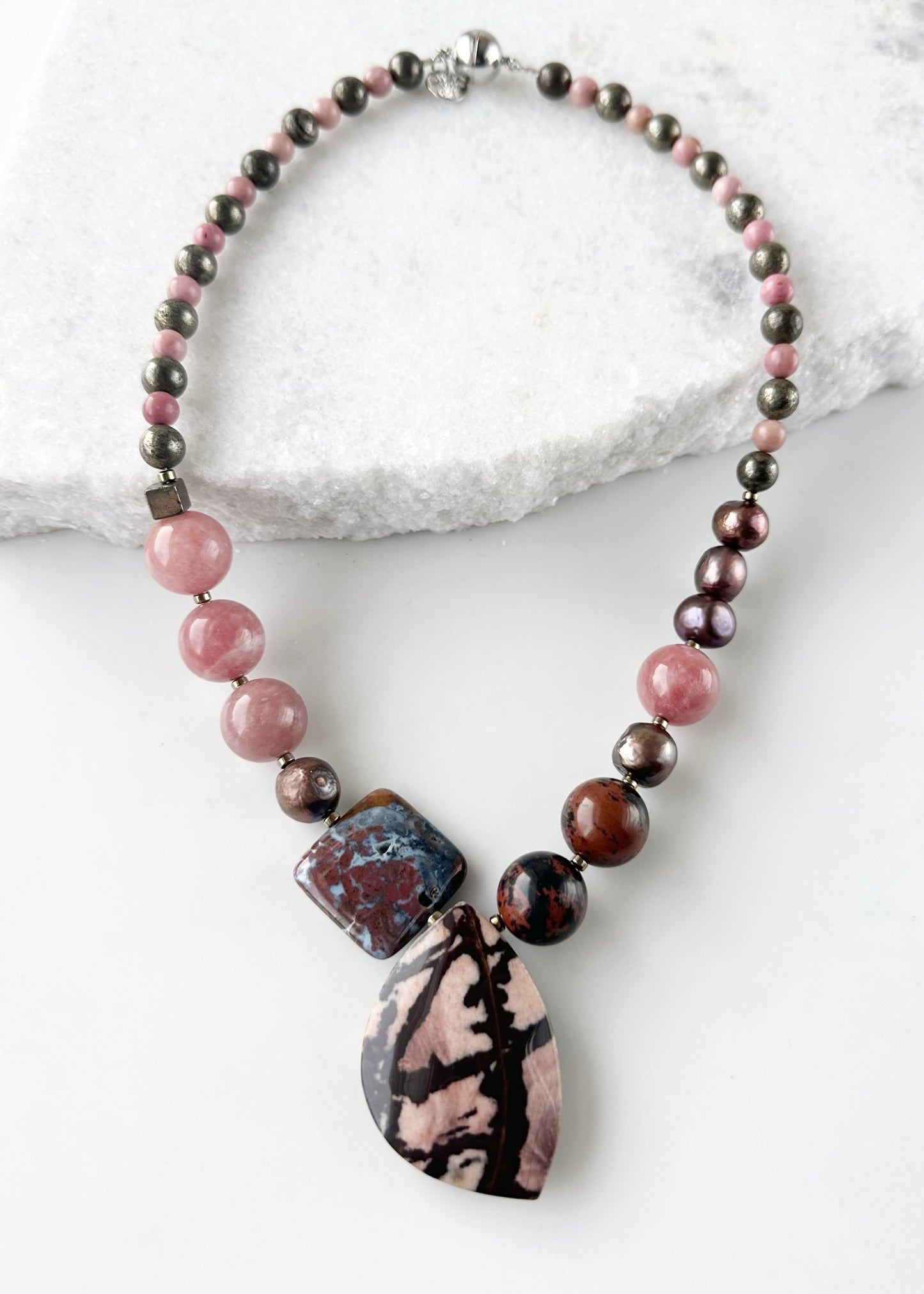 Designed and made in Canada, this Asymmetrical One-Of-A-Kind 19.5” Necklace features a beautiful mix of Stones, a Pendant of Outback Jasper, an Ocean Jasper Square, Mahogany Obsidian, Morango Quartz, Pyrite, Rhodonite and Warm Brown Pearls. This piece is finished with Gold-plated Hematite accents and a Magnetic Clasp for Secure, Easy Wear.