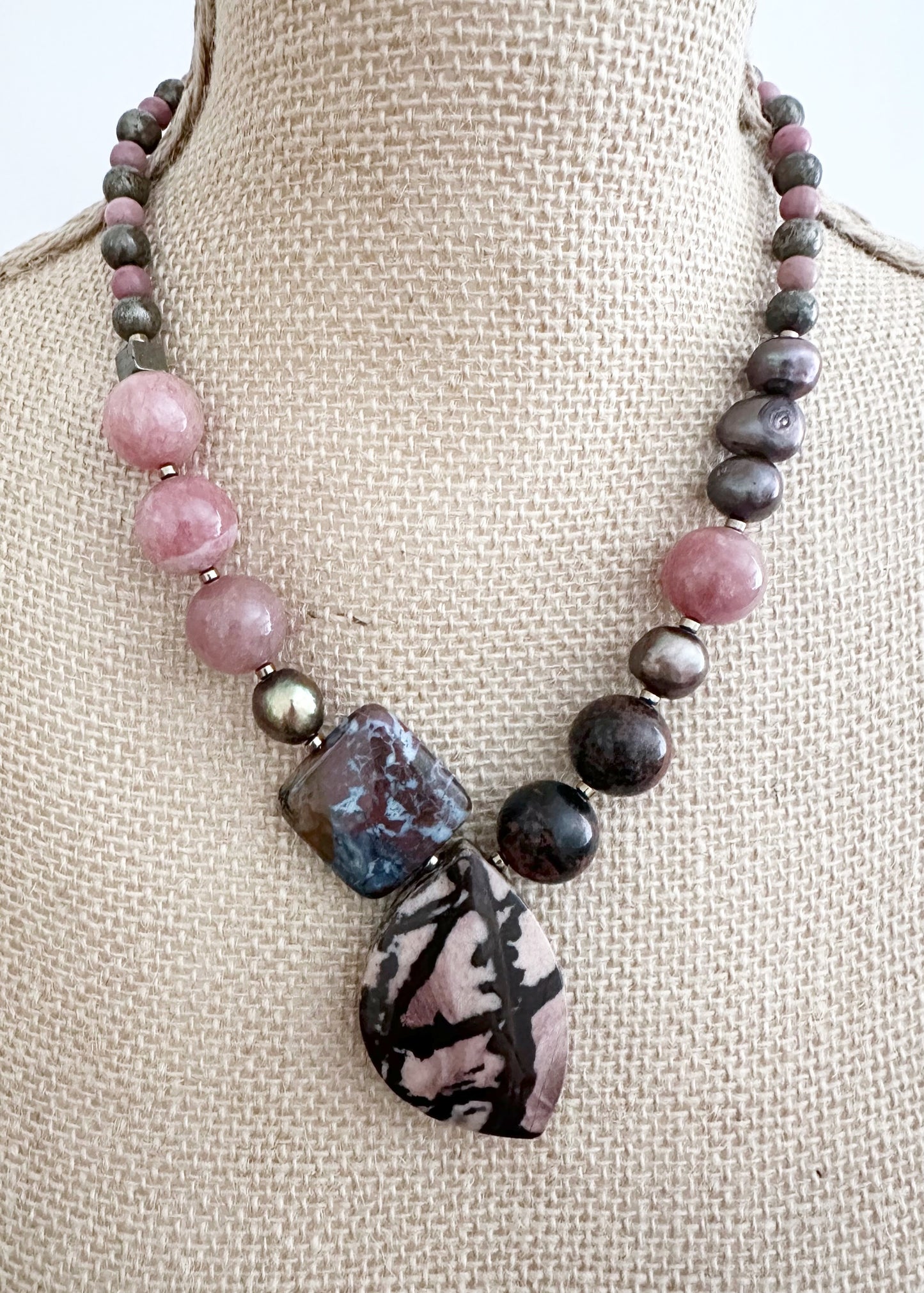 Designed and made in Canada, this Asymmetrical One-Of-A-Kind 19.5” Necklace features a beautiful mix of Stones, a Pendant of Outback Jasper, an Ocean Jasper Square, Mahogany Obsidian, Morango Quartz, Pyrite, Rhodonite and Warm Brown Pearls. This piece is finished with Gold-plated Hematite accents and a Magnetic Clasp for Secure, Easy Wear.